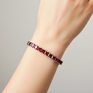 May include: A silver bracelet with a row of square-cut, deep red gemstones. The bracelet is worn on a wrist against a neutral background, showcasing the jewelry's color and design. The gemstones are evenly spaced.