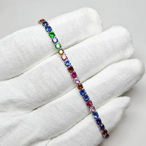 Pu&ograve; includere: Un bracciale d'argento con una fila di pietre preziose multicolori. Le pietre preziose sono di vari colori, tra cui blu, verde, giallo, rosa e rosso. Il bracciale &egrave; presentato su uno sfondo bianco.