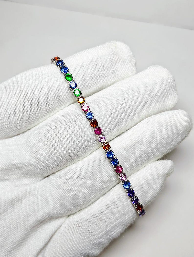 Pu&ograve; includere: Un bracciale d'argento con una fila di pietre preziose multicolori. Le pietre preziose sono di vari colori, tra cui blu, verde, giallo, rosa e rosso. Il bracciale &egrave; presentato su uno sfondo bianco.