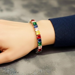 May include: A silver bracelet with a series of colorful square-cut gemstones. The gemstones are red, green, blue, yellow, and clear. The bracelet is worn on a wrist with a navy blue sleeve.