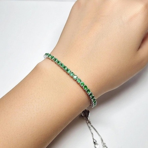Op de afbeelding: Een zilveren armband met een rij heldergroene edelstenen. De armband zit om een pols en de verstelbare sluiting hangt naar beneden. De edelstenen zijn gelijkmatig verdeeld en reflecteren licht.