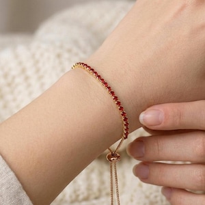 Puede incluir: Una pulsera dorada con una fila de pequeñas piedras preciosas rojas. La pulsera tiene una cadena delicada y un cierre deslizante para ajustar la talla. Las piedras preciosas están espaciadas uniformemente, creando una línea continua de color. La pulsera se lleva en la muñeca.