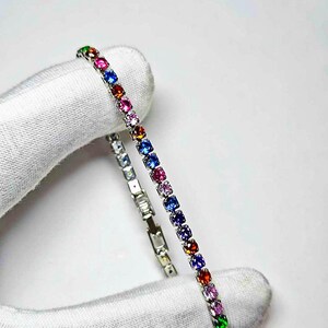 Pu&ograve; includere: Un braccialetto d'argento con una fila di pietre preziose colorate. Le pietre preziose sono in vari colori, tra cui rosso, rosa, blu, verde e viola. Il braccialetto &egrave; tenuto da una mano guantata su sfondo bianco.