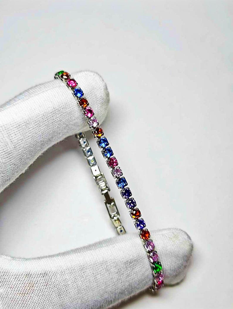 Pu&ograve; includere: Un braccialetto d'argento con una fila di pietre preziose colorate. Le pietre preziose sono in vari colori, tra cui rosso, rosa, blu, verde e viola. Il braccialetto &egrave; tenuto da una mano guantata su sfondo bianco.