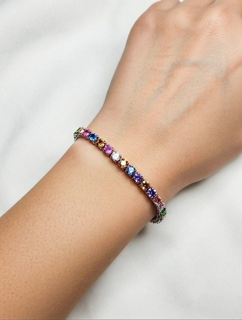 Pu&ograve; includere: Un braccialetto tennis con una fila di pietre preziose colorate. Il braccialetto presenta una variet&agrave; di colori, tra cui rosa, viola, blu, giallo e verde. Le pietre sono incastonate in una fascia d'argento o platino.