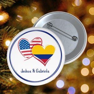 Puede incluir: Una chapa redonda con dos corazones superpuestos, uno con el patrón de la bandera de Estados Unidos y el otro con la bandera de Colombia. El texto Joshua & Gabriela está impreso en azul debajo de los corazones, dentro de un borde circular azul.