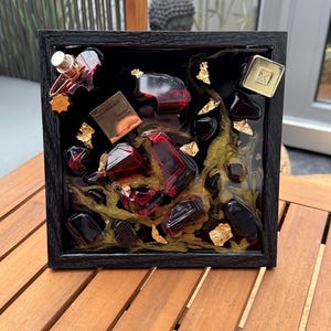 May include: A framed art piece featuring red perfume bottles, black resin, gold leaf accents, and a gold-colored square. The artwork is encased in a black wooden frame, creating a luxurious and artistic display.