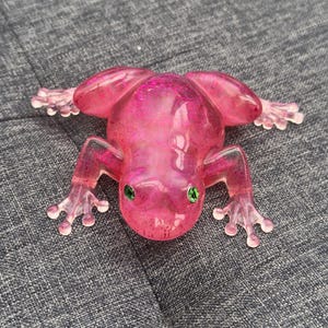 May include: A pink, translucent frog figurine with glitter accents and green eyes. The frog is posed with its legs outstretched, giving a clear view of its webbed feet. The figurine is resting on a textured gray surface.