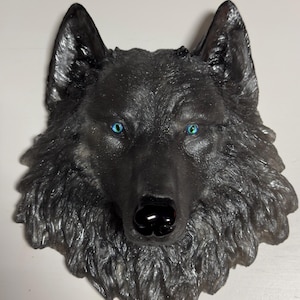 May include: A detailed, dark gray wolf head sculpture with striking blue eyes and a glossy black nose. The sculpture has a textured, fur-like appearance and is mounted on a white surface. The artwork is a decorative piece.