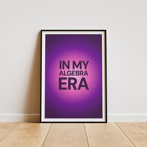 May include: A framed poster with the text "IN MY ALGEBRA ERA" in dark purple against a gradient purple and pink background. The poster is in a black frame and leans against a white wall on a wooden floor.
