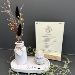 Könnte beinhalten: Eine weiße Keramikvase mit Trockenblumen und einer kleinen Zwergfigur auf einem weißen Tablett. Auf dem Tablett steht das Wort "Schön". Im Hintergrund befindet sich eine Karte mit Text.