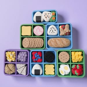 May include: A collection of seven square and rectangular bento-style containers filled with miniature food-shaped erasers. The set includes various items such as rice balls, bread, cookies, fruits, vegetables, cheese, and eggs in vibrant colors.