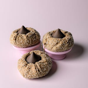 May include: Three brown, round, baked goods with a dark chocolate center, sprinkled with white sugar crystals, sit on small pink pedestals against a pink background. A small chain is attached to one of the baked goods.