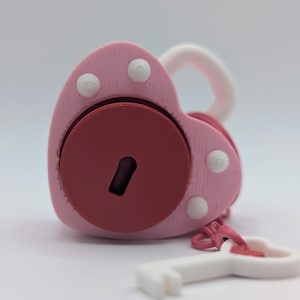 May include: A pink heart-shaped padlock with a red circular center and a white key. The padlock has a white handle and three white circular accents. The key is white with a pink heart-shaped head.