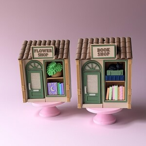 May include: Two miniature shop models, one labeled "Flower Shop" and the other "Book Shop." The flower shop has a green door and a plant. The book shop has a black cat and books. Both are on pink pedestals.