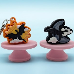 May include: Two silicone charms featuring black dragon silhouettes. The left charm is orange with a yellow sunburst and white clouds, including a metal keychain ring. The right charm is black with a grey crescent moon and white clouds.