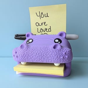 May include: A purple hippo-shaped desk organizer holding a stack of yellow sticky notes. The notes have the handwritten message "You are Loved". A black pen and a white pen are inserted into the top of the hippo.