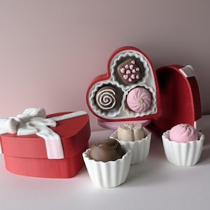 May include: A red heart-shaped box with a white bow and several chocolates. The chocolates are in white cupcake-shaped holders. Some chocolates are in a heart-shaped box with a white interior. The chocolates are various colors and designs.