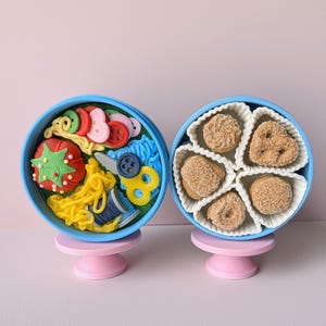 May include: Two blue, round containers on pink pedestals. One holds sewing notions: a red tomato, yellow thread, blue thread spool, scissors, and buttons. The other contains five small, round, and heart-shaped pastries.