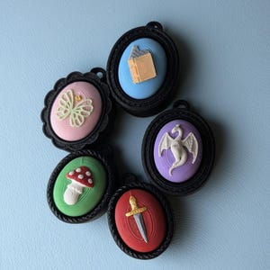May include: Five oval-shaped pendants with black frames. Each pendant features a different design: a butterfly, a mushroom, a dragon, a book, and a sword. The backgrounds are various colors, including pink, green, blue, and red.