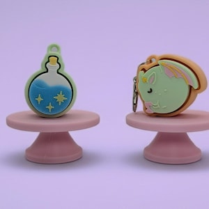 May include: Two silicone charms on pink stands. One charm is a round light green potion bottle with blue liquid and yellow star accents. The other charm is a light green winged creature with pink wing details and a tan border, featuring a metal ring.