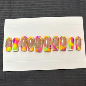 Pode incluir: Um conjunto de dez unhas artificiais com um design tie-dye em tons de rosa, amarelo e verde. Cada unha apresenta um centro castanho e embelezamentos dourados. As unhas são apresentadas num cartão branco.