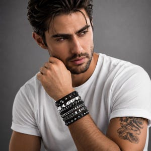 May include: A stack of black leather bracelets with silver and black metal accents. The bracelets include pyramid studs, chain links, and smooth bands. The bracelets are worn on a person's wrist, against a white t-shirt.