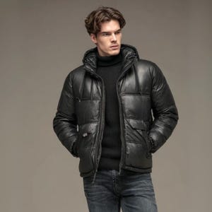 May include: A black puffer jacket with a hood, worn over a black turtleneck sweater. The jacket has a quilted design and two front pockets. The model is wearing blue jeans.