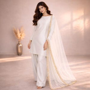 May include: A white, long-sleeved tunic and wide-leg pants ensemble. The outfit includes a sheer, flowing scarf with gold trim. The model is wearing gold earrings and sandals. The background is a soft, neutral color.