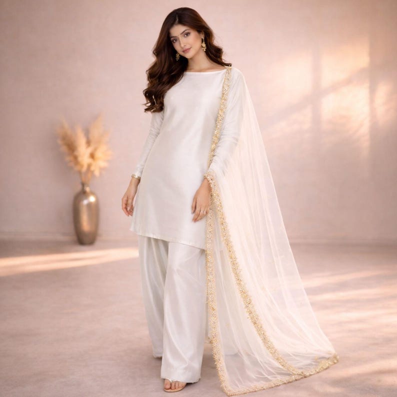 May include: A white, long-sleeved tunic and wide-leg pants ensemble. The outfit includes a sheer, flowing scarf with gold trim. The model is wearing gold earrings and sandals. The background is a soft, neutral color.