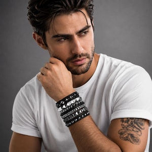 May include: A stack of black leather bracelets with silver and black metal accents. The bracelets include pyramid studs, chain links, and smooth bands. The bracelets are worn on a person's wrist, against a white t-shirt.