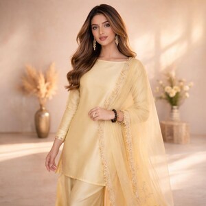 May include: A light yellow, long-sleeved tunic and matching pants with a sheer scarf. The tunic has a boat neck and is trimmed with gold embroidery. The outfit is part of a traditional South Asian ensemble.