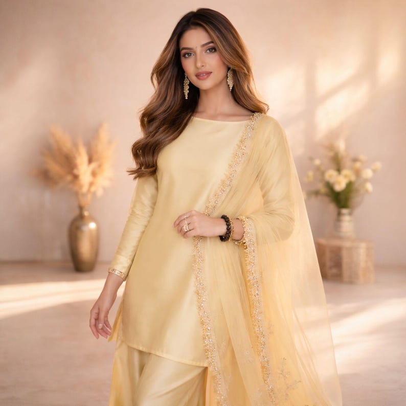 May include: A light yellow, long-sleeved tunic and matching pants with a sheer scarf. The tunic has a boat neck and is trimmed with gold embroidery. The outfit is part of a traditional South Asian ensemble.