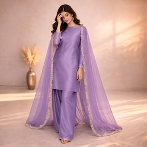 May include: A lilac-colored, two-piece outfit featuring a long-sleeved tunic and wide-leg pants. A sheer, matching cape with gold trim drapes over the shoulders. The outfit is made of a smooth, shiny fabric, and the overall style is elegant.
