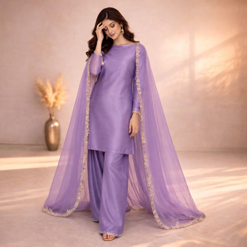 May include: A lilac-colored, two-piece outfit featuring a long-sleeved tunic and wide-leg pants. A sheer, matching cape with gold trim drapes over the shoulders. The outfit is made of a smooth, shiny fabric, and the overall style is elegant.