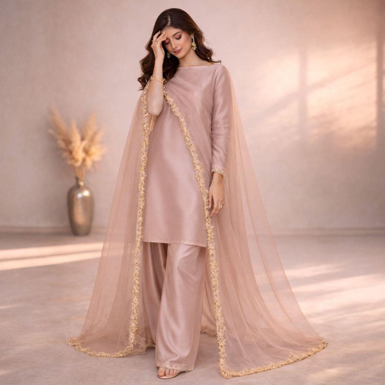 May include: A blush pink ensemble featuring a long-sleeved tunic and wide-leg pants. A sheer, matching cape with gold trim drapes over the shoulders. The outfit is made of a silky material, and the overall style is elegant.