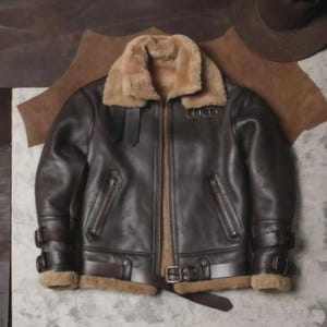 Pode incluir: Casaco de couro castanho com gola e forro de pele de carneiro. O casaco apresenta fecho de correr, dois bolsos frontais e correias ajustáveis nas mangas e na cintura. Estilo aviador clássico.