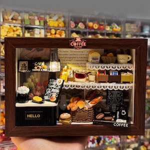 May include: A miniature diorama of a coffee shop inside a dark brown wooden frame. The scene features shelves with tiny baskets, bread, pastries, and a sign that reads COFFEE. A counter displays a sign saying HELLO, a black coffee machine, and various food items.