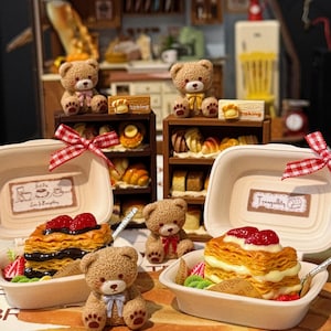 May include: Miniature bakery scene featuring teddy bears and pastries. Two open containers display layered desserts with strawberries. Miniature shelves hold various baked goods. The scene includes a small kitchen setting with a refrigerator and a bread loaf.