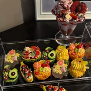 May include: A collection of miniature dessert sculptures, including sundaes, parfaits, and fruit-topped treats. The desserts feature a variety of fruits, whipped cream, and chocolate. The display includes a large parfait and several smaller, individual desserts.