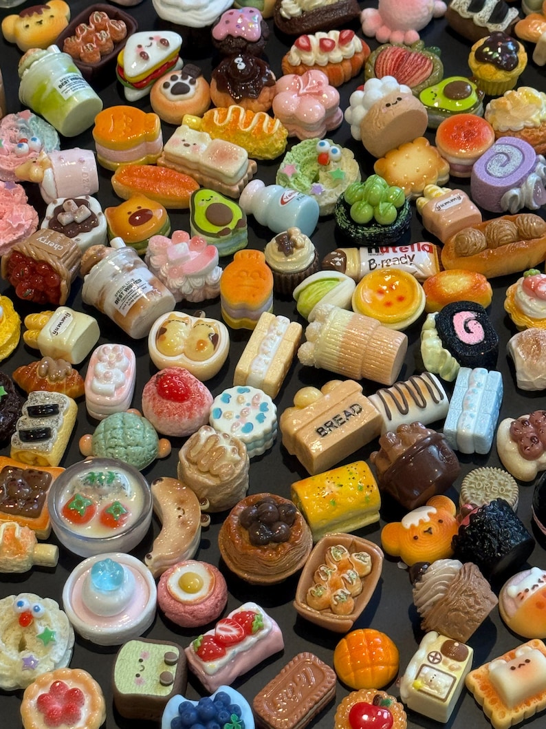 May include: A collection of miniature food charms in various shapes and colors. The charms include tiny cakes, pastries, bread, and other dessert-themed items. Some charms have text like "Bread" and "Nutella B-ready".