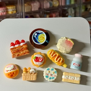 May include: A collection of miniature food items, including a coffee cup with a swirling design, pastries with strawberries and cream, a corn dog, and a small bottle of Yakult Plus. The items are brightly colored and detailed.