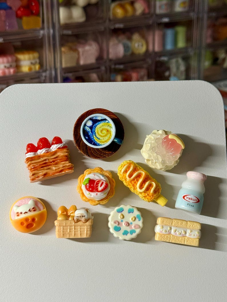 May include: A collection of miniature food items, including a coffee cup with a swirling design, pastries with strawberries and cream, a corn dog, and a small bottle of Yakult Plus. The items are brightly colored and detailed.