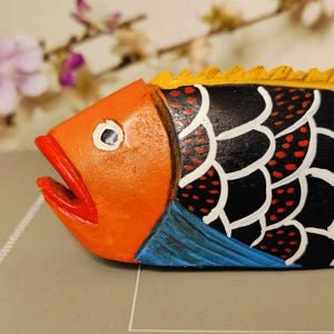 May include: A hand-painted wooden fish sculpture. The fish features an orange head with a red mouth, black body with white and red accents, yellow dorsal fin, and blue fins. The sculpture is a decorative item.