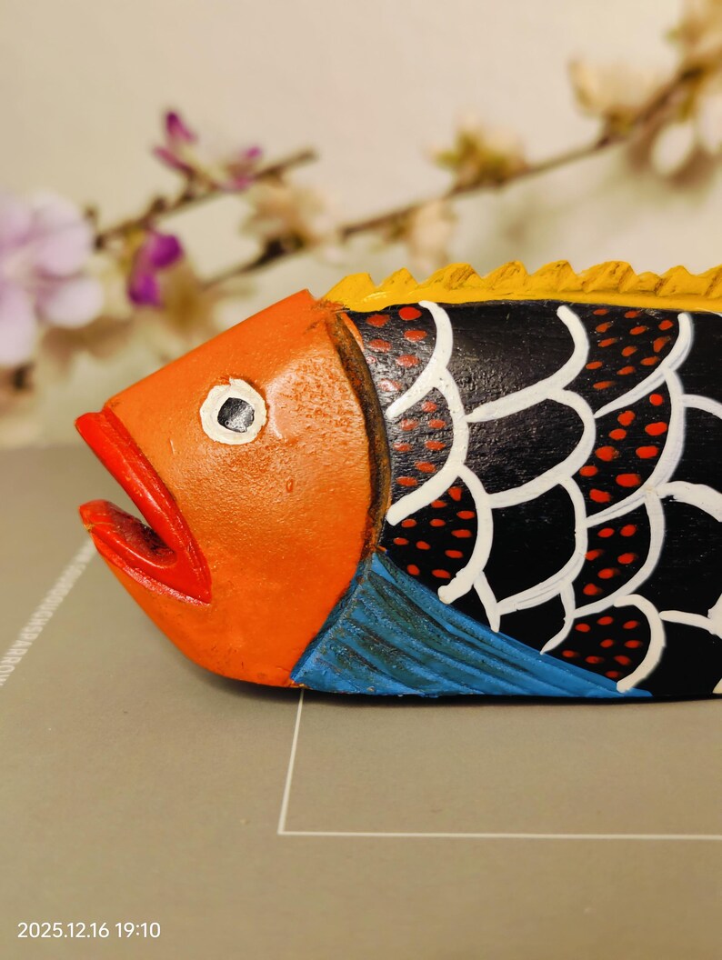 May include: A hand-painted wooden fish sculpture. The fish features an orange head with a red mouth, black body with white and red accents, yellow dorsal fin, and blue fins. The sculpture is a decorative item.