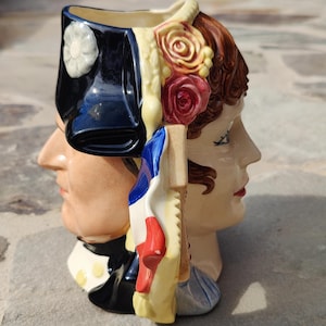 Puede incluir: Taza de cerámica con dos caras, un hombre y una mujer, de espaldas. El lado masculino tiene un sombrero azul oscuro con una flor blanca, mientras que el lado femenino tiene cabello castaño con rosas. Un diseño de bandera francesa está en el lateral.