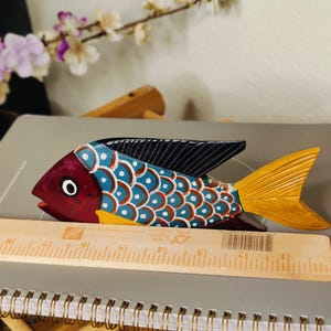May include: A hand-painted wooden fish sculpture with a red head, blue and white body, and yellow tail. The fish is approximately 10 inches long and has black fins. It is resting on a notebook with a wooden ruler.