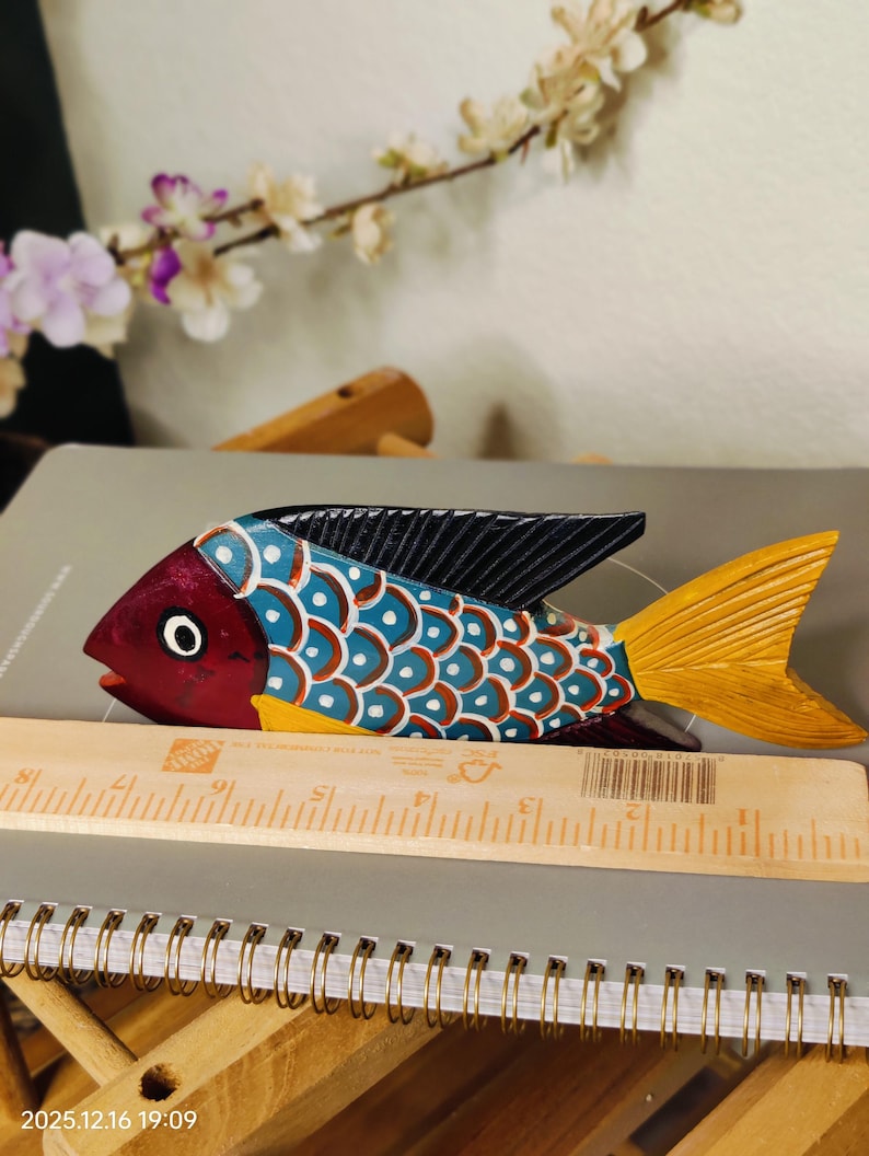 May include: A hand-painted wooden fish sculpture with a red head, blue and white body, and yellow tail. The fish is approximately 10 inches long and has black fins. It is resting on a notebook with a wooden ruler.