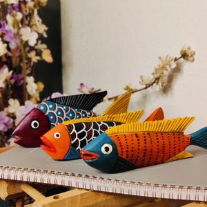 May include: Three colorful wooden fish sculptures. The fish are painted in shades of red, orange, and blue, with intricate patterns and details. The fish are arranged on a notebook, with a wooden stand in the foreground and flowers in the background.