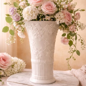 Puede incluir: Un jarrón blanco y ornamentado lleno de un exuberante arreglo de rosas rosas, hortensias blancas y delicada vegetación. El jarrón se encuentra sobre una bandeja redonda con tapa de mármol, creando una estética suave y romántica.